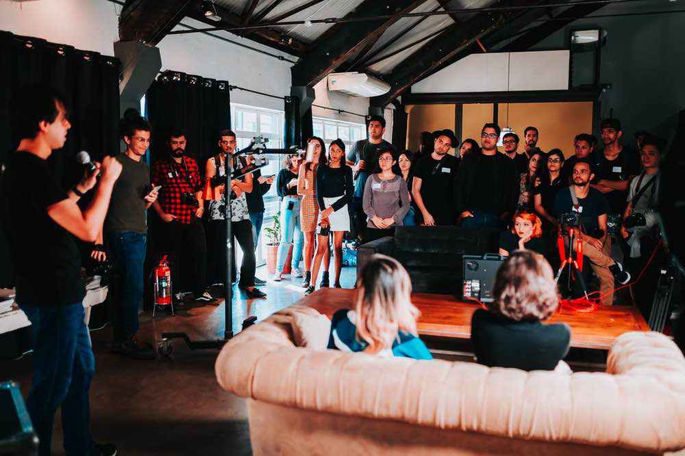 A speaker holding a microphone faces a diverse group of seated and standing attendees in a bright industrial loft space.