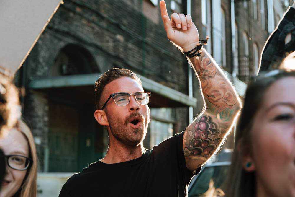 A young man with glasses and tattooed arm raises his fist enthusiastically on a city street, fellow pedestrians in soft focus behind him.