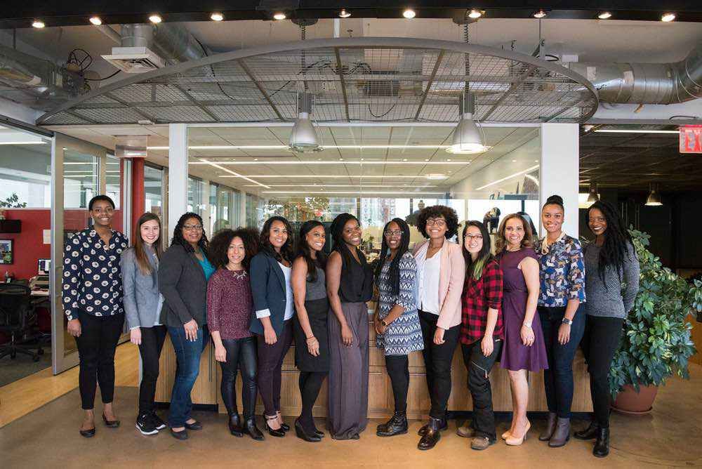 Thirteen women of various backgrounds stand side by side in a modern open-plan office, smiling toward the camera.