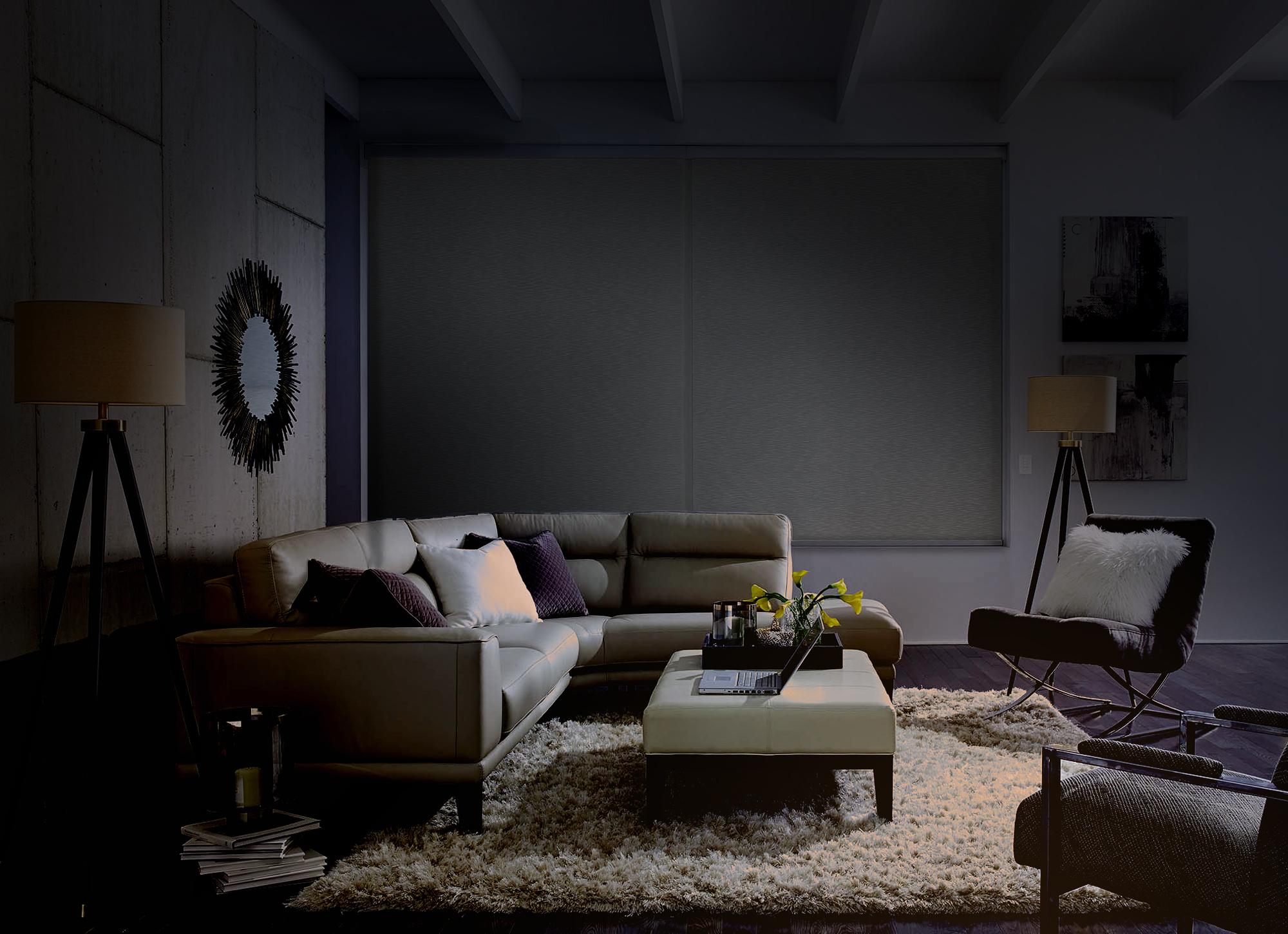 A dimly lit living room at night, with motorized shades fully lowered over a large window; the room’s furnishings—a sectional sofa, ottoman, floor lamp, and accent chair—are barely visible in the low light, creating a cozy cinema-like atmosphere.