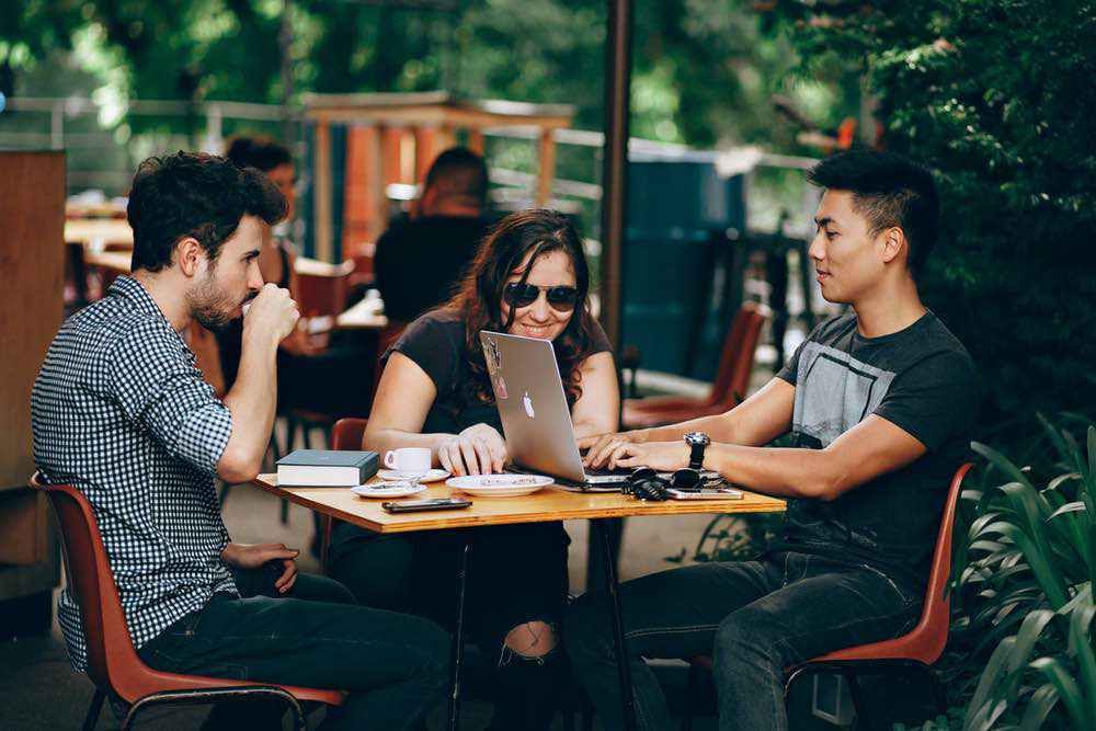 Three colleagues sit around a wooden café table—one sipping coffee, another smiling at a laptop screen, and a third typing—beneath a canopy of greenery.