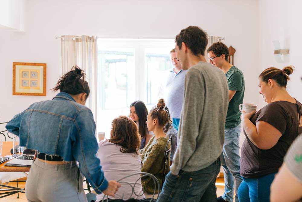 A mixed group of six people lean over laptops on a long dining table in a sunlit room, collaborating on a project.