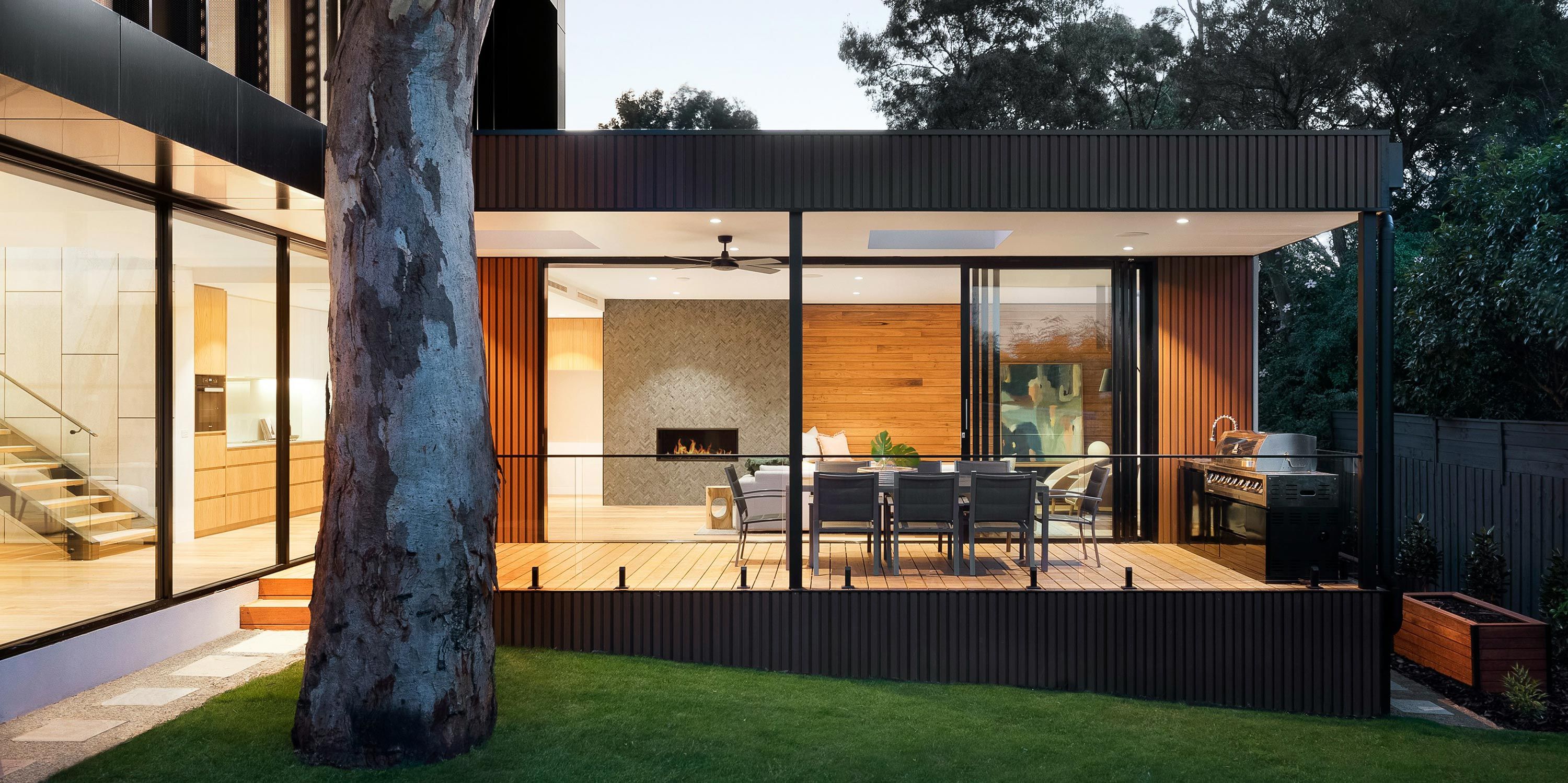 A stylish outdoor living area with a dining table, barbecue grill, and a ceiling fan, enclosed by glass walls and surrounded by greenery.